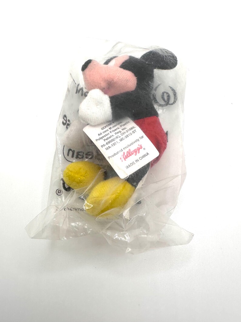 Pu&ograve; includere: Un piccolo peluche di Topolino in un sacchetto di plastica trasparente. Il giocattolo ha dettagli neri, rossi, bianchi, rosa e gialli. Un'etichetta con testo &egrave; attaccata. La confezione include il logo Kellogg's.