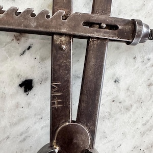May include: A vintage metal caliper tool featuring a dark, weathered finish with a serrated adjustment bar and a central pivot point. The metal surface displays visible signs of age and wear, and the letters HM are clearly stamped into the metal arm.