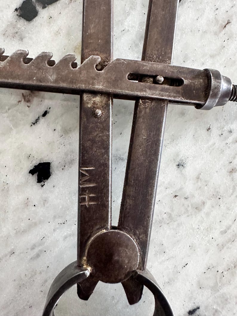 May include: A vintage metal caliper tool featuring a dark, weathered finish with a serrated adjustment bar and a central pivot point. The metal surface displays visible signs of age and wear, and the letters HM are clearly stamped into the metal arm.
