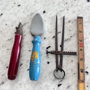 May include: A set of three tools including a red-handled scribe, a blue-handled sharpening stone with the text 'Knife Sharp No 275 A', and a metal caliper, positioned next to a wooden ruler measuring up to 9 inches in length.