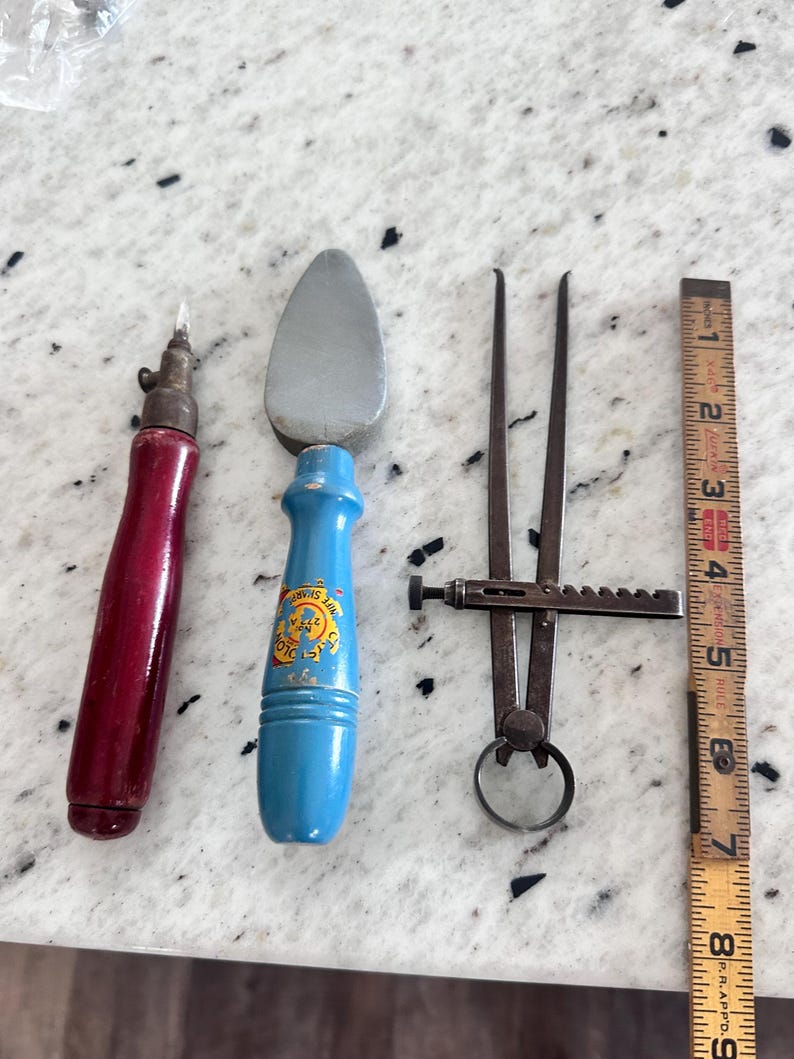 May include: A set of three tools including a red-handled scribe, a blue-handled sharpening stone with the text 'Knife Sharp No 275 A', and a metal caliper, positioned next to a wooden ruler measuring up to 9 inches in length.