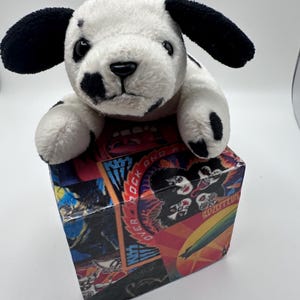 May include: A plush white dog with black ears and spots resting on a square box decorated with colorful rock music graphics, including the band name KISS and a Led Zeppelin airship illustration, featuring vibrant red, blue, and yellow tones.