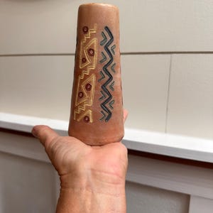 May include: A terracotta-colored vase with geometric patterns in gold, black, and teal. The vase has a tapered shape and is held in a hand. The design features zigzags and angular shapes, suggesting an ethnic or tribal influence.