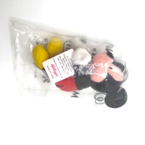 Pu&ograve; includere: Un peluche di Topolino, confezionato in un sacchetto di plastica trasparente. Il giocattolo presenta orecchie nere, un fiocco a pois rossi e bianchi e piedi gialli. Un'etichetta Kellogg's &egrave; attaccata alla confezione.