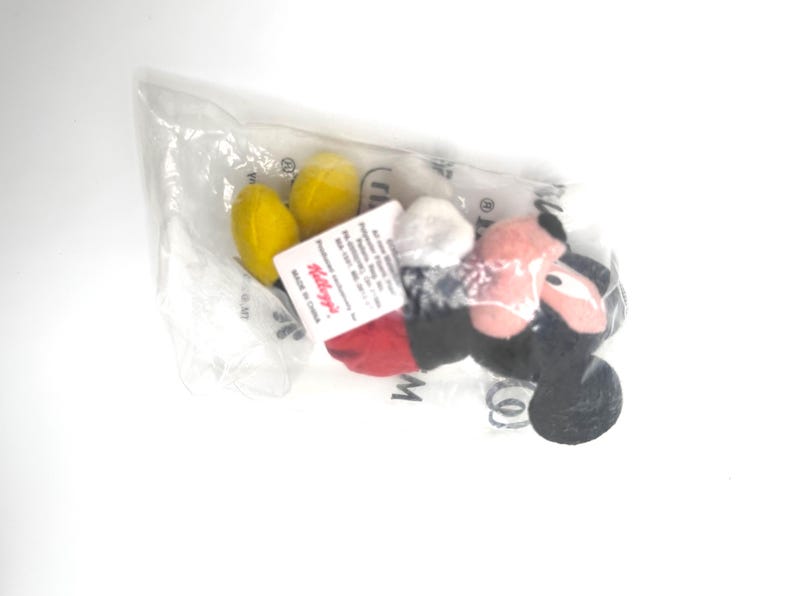 Pu&ograve; includere: Un peluche di Topolino, confezionato in un sacchetto di plastica trasparente. Il giocattolo presenta orecchie nere, un fiocco a pois rossi e bianchi e piedi gialli. Un'etichetta Kellogg's &egrave; attaccata alla confezione.