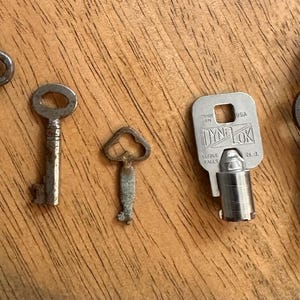 May include: Five antique keys of varying sizes and designs are arranged on a wooden surface. The keys are made of metal, with a range of shapes, including a decorative floral design, a simple oval, and a key with the text "DYNA LOCK" on it.