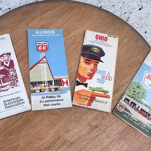 May include: Four vintage travel brochures on a wooden surface. The brochures feature text and illustrations for Eastern States South, Illinois Phillips 66, Ohio, and Kentucky Tennessee Sinclair. The brochures are in various colors.