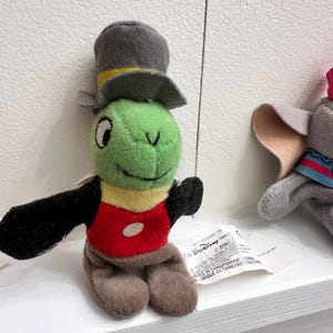 May include: A plush Jiminy Cricket toy featuring a green head, a grey top hat with a yellow band, a red vest with a white circular button, black arms, and brown legs. A white fabric tag with The Walt Disney Company text is attached to the side.