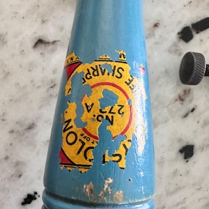 May include: A light blue wooden handle with a weathered yellow, red, and black circular label featuring the text "COLON" and "NO: 272 A". The label shows significant wear and peeling, revealing the blue paint underneath on the cylindrical surface.