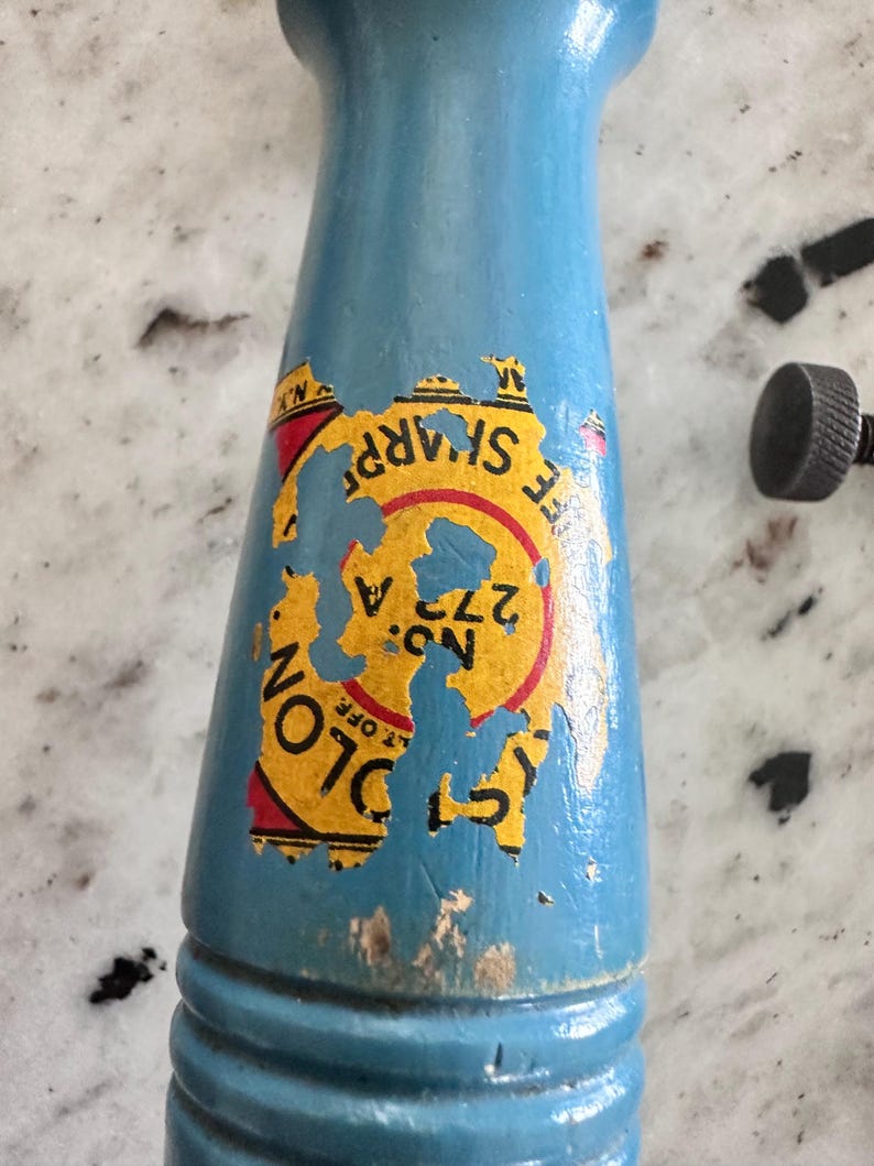 May include: A light blue wooden handle with a weathered yellow, red, and black circular label featuring the text "COLON" and "NO: 272 A". The label shows significant wear and peeling, revealing the blue paint underneath on the cylindrical surface.