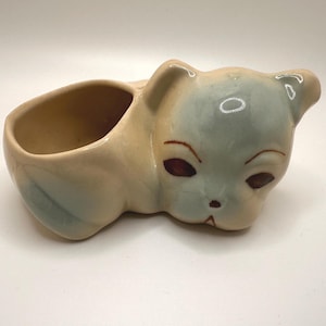 May include: A vintage ceramic cat planter in a reclining pose. The planter is primarily beige with light blue accents on the head and tail. The cat's eyes are a deep maroon color. The planter has a single opening for plants.