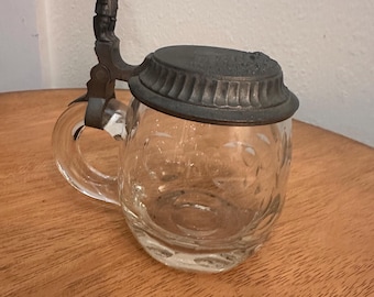 Op de afbeelding: Heldere glazen bierpul met een decoratief metalen deksel en handvat. Het deksel is donkergrijs en het handvat heeft een klein figuur. Het glas heeft een geëtst bloemmotief. De pul staat op een houten oppervlak.