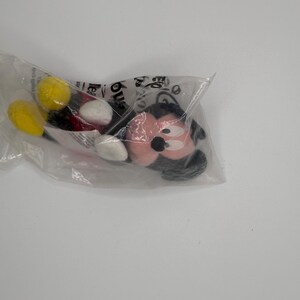 Pu&ograve; includere: Un peluche di Topolino in un sacchetto di plastica trasparente. Il peluche presenta un viso rosa, orecchie nere e dettagli in bianco e nero. Sono visibili le gambe gialle e nere del giocattolo.