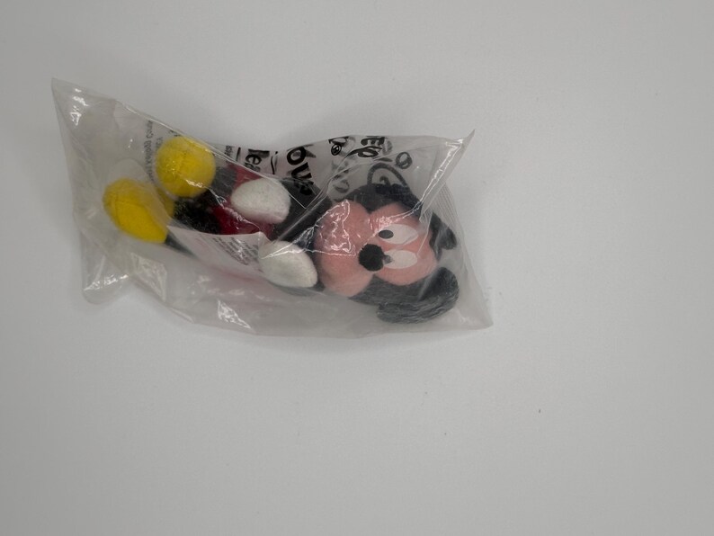 Pu&ograve; includere: Un peluche di Topolino in un sacchetto di plastica trasparente. Il peluche presenta un viso rosa, orecchie nere e dettagli in bianco e nero. Sono visibili le gambe gialle e nere del giocattolo.