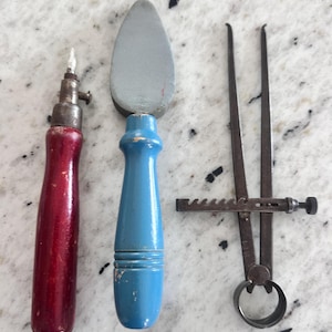 May include: A set of three tools including a red-handled metal awl, a blue-handled gray stone burnisher, and a dark metal caliper with a serrated adjustment bar. Each tool features a distinct handle or mechanism for manual craft and measurement tasks.