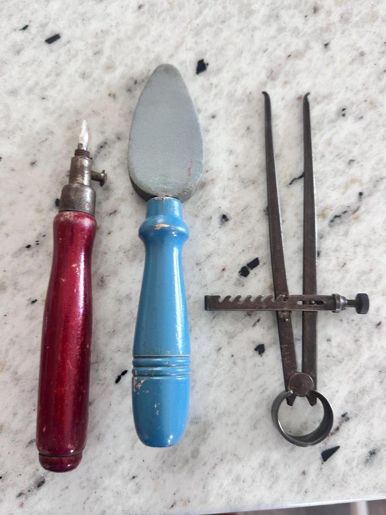 May include: A set of three tools including a red-handled metal awl, a blue-handled gray stone burnisher, and a dark metal caliper with a serrated adjustment bar. Each tool features a distinct handle or mechanism for manual craft and measurement tasks.