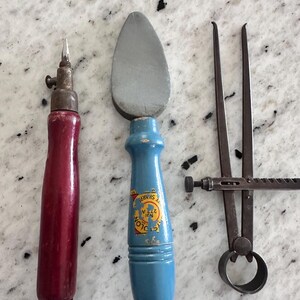 May include: Three vintage hand tools: a red wooden-handled scribe with a metal tip, a blue wooden-handled tool with a grey stone head featuring a yellow label that reads "COLON" and "SHARP", and a dark metal caliper with a circular base.