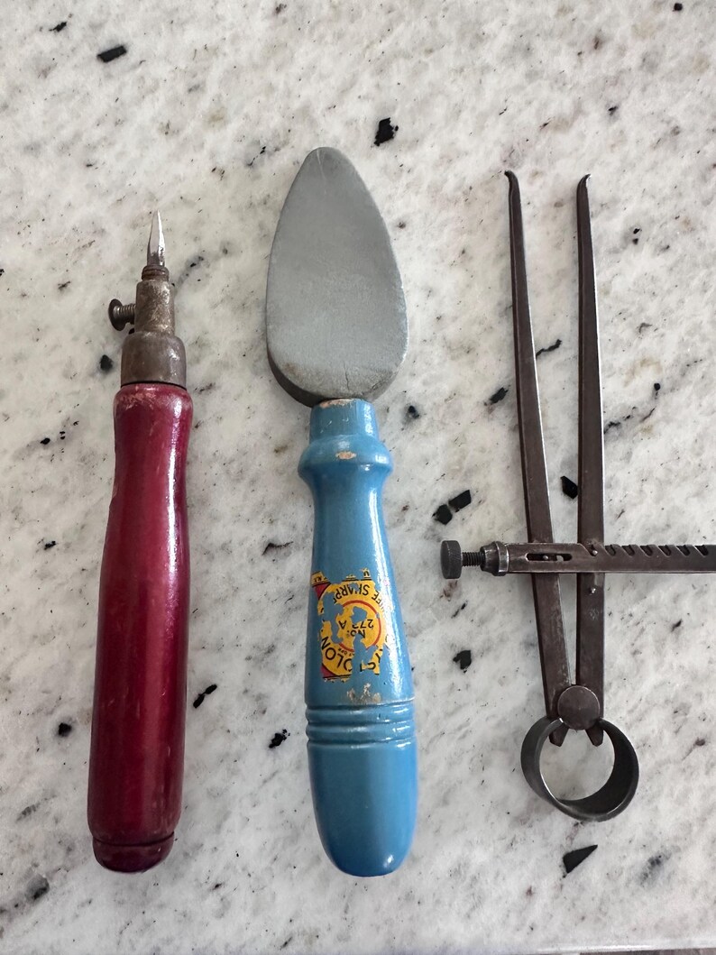 May include: Three vintage hand tools: a red wooden-handled scribe with a metal tip, a blue wooden-handled tool with a grey stone head featuring a yellow label that reads "COLON" and "SHARP", and a dark metal caliper with a circular base.