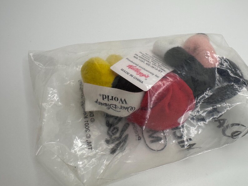Pu&ograve; includere: Un peluche di Topolino in un sacchetto di plastica trasparente. Il giocattolo presenta mani gialle, guanti neri, pantaloncini rossi e viso bianco e rosa. La borsa contiene etichette con testo, tra cui "Walt Disney World" e "Kellogg's".