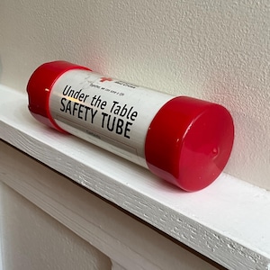 May include: A cylindrical safety tube with red caps and a white body, labeled "Under the Table SAFETY TUBE." The Red Cross logo is visible. The tube rests on a white shelf against a neutral wall.