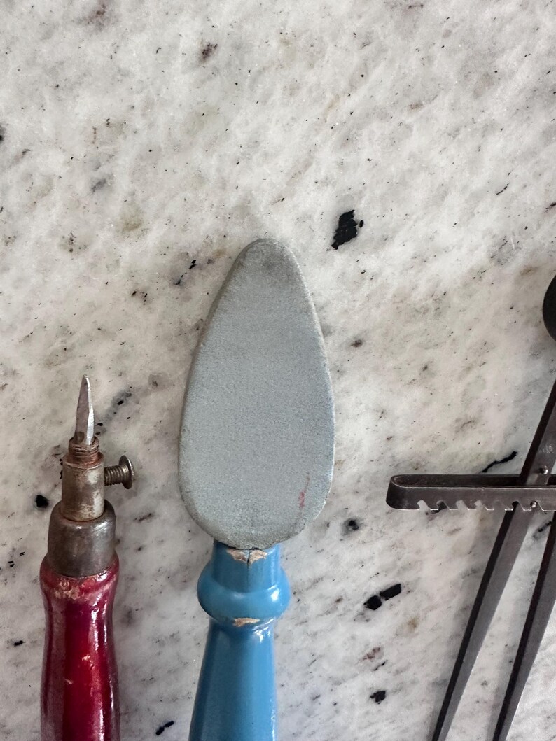 May include: A tool with a pointed metal tip and a red wooden handle, a tool with a gray, teardrop-shaped abrasive head and a blue wooden handle, and a dark metal tool with a serrated edge and a long, thin, straight metal arm.