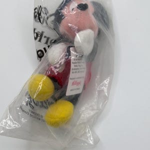 Pu&ograve; includere: Un peluche di Topolino in un sacchetto di plastica trasparente. Il peluche ha una camicia rossa, scarpe gialle e orecchie nere. La borsa ha testo e un logo Disney.