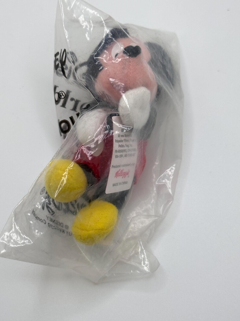 Pu&ograve; includere: Un peluche di Topolino in un sacchetto di plastica trasparente. Il peluche ha una camicia rossa, scarpe gialle e orecchie nere. La borsa ha testo e un logo Disney.