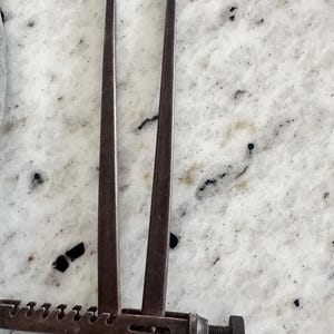 May include: A dark gray metal caliper tool featuring two long, slender, slightly curved arms connected by a horizontal bar with a series of notches and a threaded adjustment knob at the end for precise measurement and spacing control.