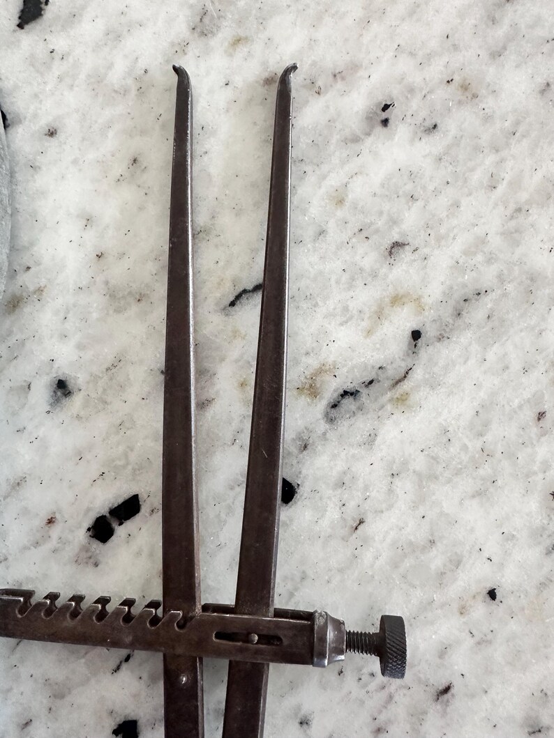 May include: A dark gray metal caliper tool featuring two long, slender, slightly curved arms connected by a horizontal bar with a series of notches and a threaded adjustment knob at the end for precise measurement and spacing control.