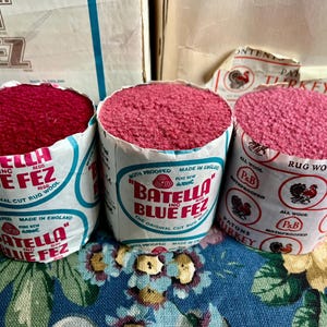 Peut inclure: Trois bobines cylindriques de laine pour tapis, dans des tons de rouge et de rose, étiquetées "Batella Blue Fez" et "Patons Turkey Rug Wool". Les bobines sont enveloppées de papier avec des graphiques et du texte vintage, sur un fond à motifs floraux.