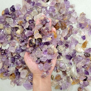 May include: A hand holding a pile of amethyst crystals. The crystals are various shades of purple and clear, and some have a golden hue.