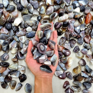 May include: A collection of gray, brown, and white agate tumbled stones. The stones are smooth and polished, with a variety of patterns and colors. The stones are arranged in a pile on a white surface.