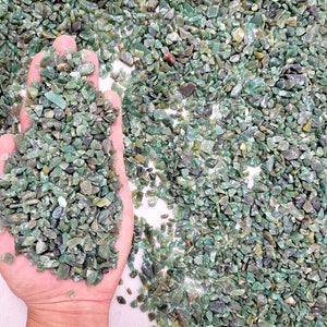 May include: A close-up of a hand holding a pile of small, green, and translucent gemstone chips. The chips are scattered on a white surface.