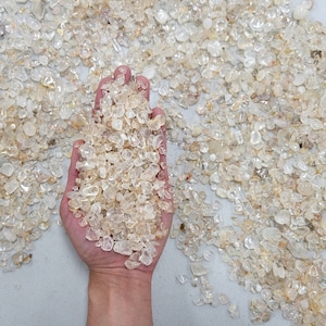 May include: A hand holding a pile of small, clear quartz crystals. The crystals are various shapes and sizes, and they have a slightly yellow tint.