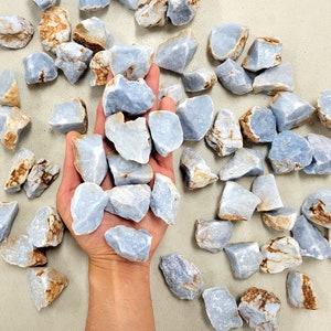 May include: A collection of light blue celestite crystal stones. The stones are rough and have a natural, earthy look. The stones are arranged in a hand and scattered on a light beige surface.