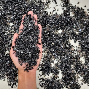 May include: A hand holding a pile of black obsidian chips. The chips are small and irregular in shape, and they have a shiny, almost metallic finish.