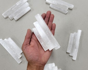 May include: A hand holding several white selenite sticks, with more sticks scattered on a gray surface.