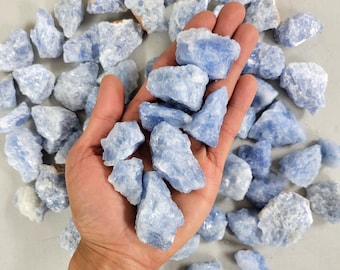 May include: A collection of light blue rough-cut crystals, with a handful held in a person's open palm. The crystals have a natural, textured appearance and vary in size. The background is a neutral, light gray.