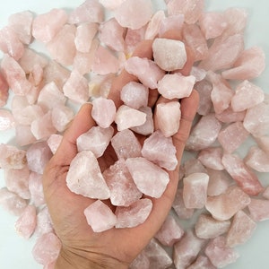May include: A handful of rough, light pink rose quartz stones. The stones vary in size and shape, with a pale pink hue. The stones are held in an open hand, with many more scattered around.