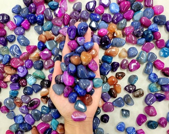 Puede incluir: Una mano sosteniendo una variedad de piedras de ágata de colores. Las piedras son lisas y pulidas, y vienen en tonos de morado, azul, rosa y marrón.