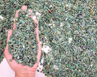 May include: A close-up of a hand holding a pile of small, green, and translucent gemstone chips. The chips are scattered on a white surface.