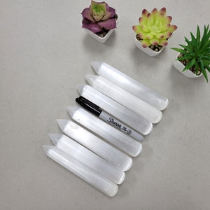 May include: Seven white selenite wands with pointed ends arranged in a fan shape. A black Sharpie marker is lying on top of the wands.