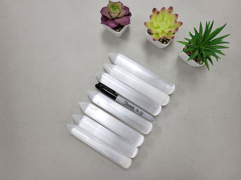 May include: Seven white selenite wands with pointed ends arranged in a fan shape. A black Sharpie marker is lying on top of the wands.