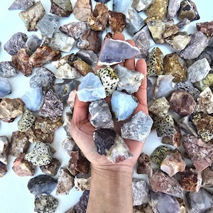 May include: A collection of rough, natural, colorful, and patterned stones. The stones are various shades of brown, gray, white, and blue. Some stones have black speckles. The stones are arranged on a white surface.