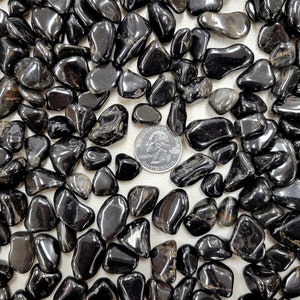 Puede incluir: Primer plano de una colecci&oacute;n de piedras de obsidiana negra pulidas. Las piedras son lisas y pulidas, y est&aacute;n dispuestas en un patr&oacute;n aleatorio. Una moneda de 2 euros se incluye para la escala.