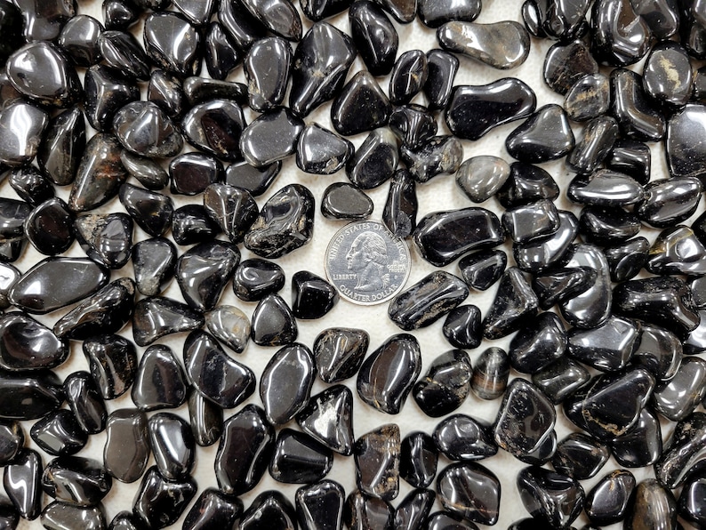 Puede incluir: Primer plano de una colecci&oacute;n de piedras de obsidiana negra pulidas. Las piedras son lisas y pulidas, y est&aacute;n dispuestas en un patr&oacute;n aleatorio. Una moneda de 2 euros se incluye para la escala.