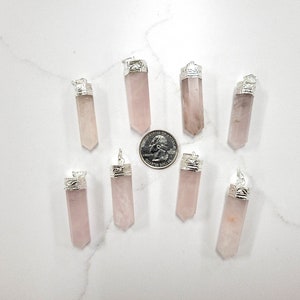 May include: Ten pink rose quartz crystal pendants with silver caps. The pendants are arranged in a circle on a white surface.