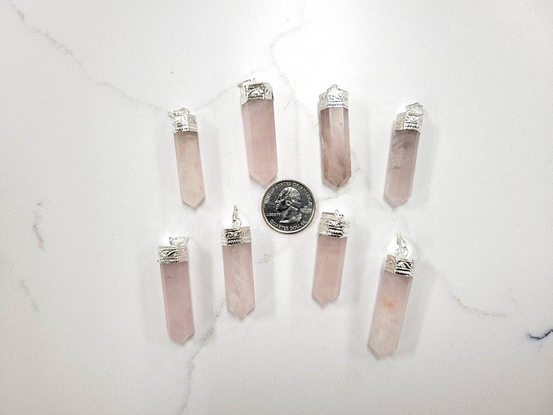 May include: Ten pink rose quartz crystal pendants with silver caps. The pendants are arranged in a circle on a white surface.