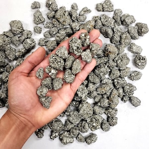 May include: A large pile of small, rough, gold-colored pyrite stones. The stones are scattered on a white surface. A hand is holding a handful of the stones.