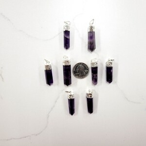 May include: Eight amethyst crystal pendants with silver caps. The pendants are arranged in a star shape on a white surface. A US quarter is included for size reference.